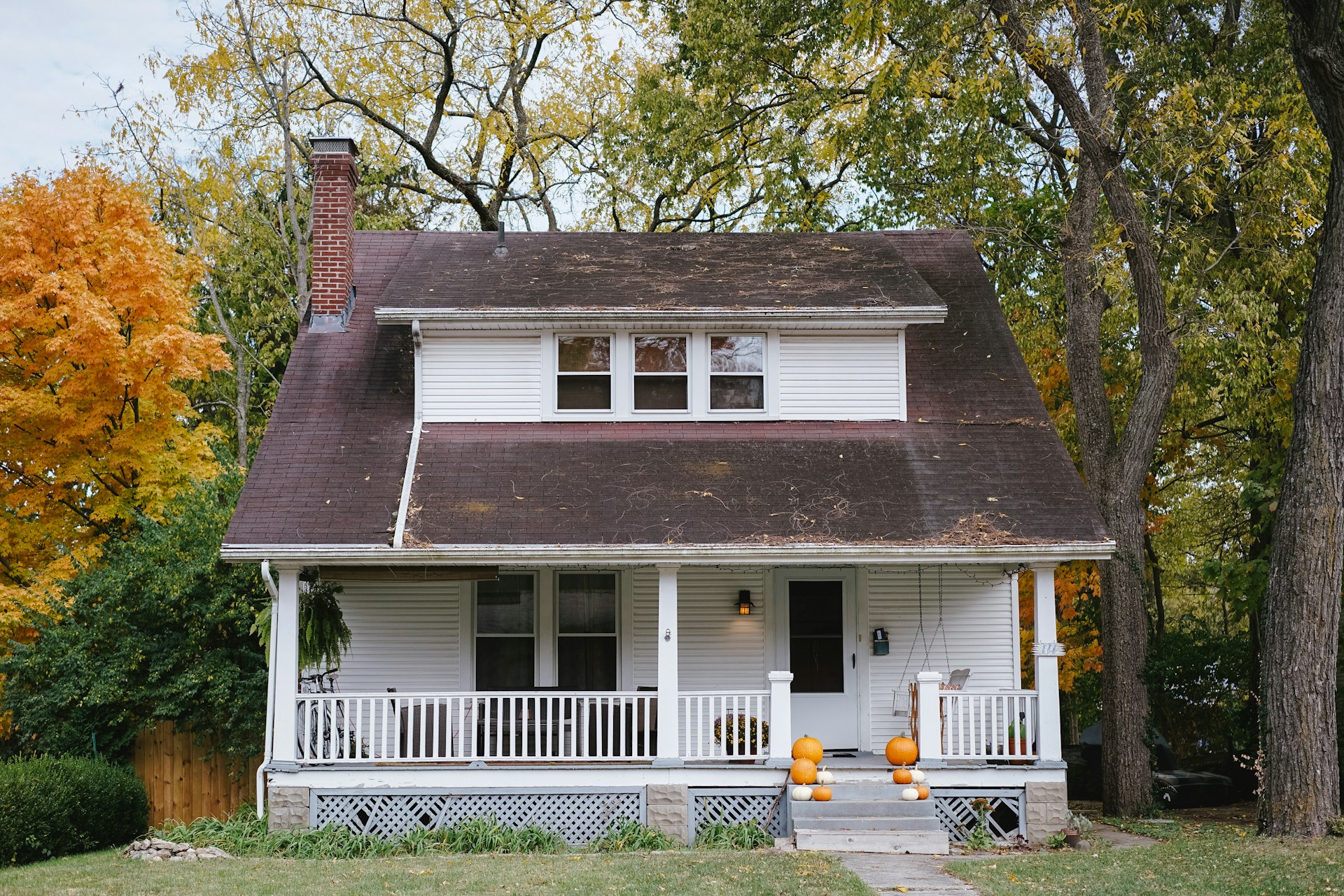 A perfectly fine house in need of some simple maintenance, such as new shingles and repaired lattice work. The photo appears to have been taken in autumn, with pumpkins lining the porch steps.